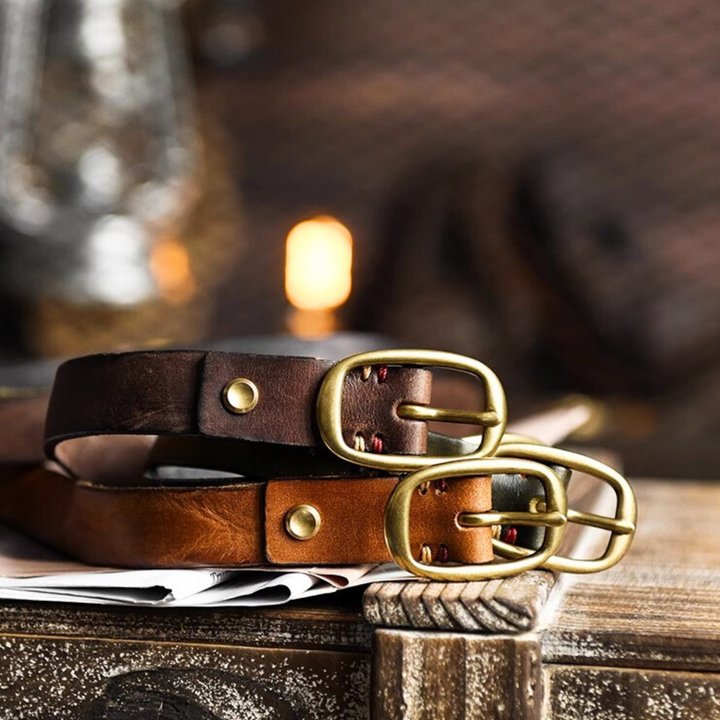 Two leather belts, one dark brown and one light brown, with brass buckles, rest on a wooden surface with papers beneath them—a perfect touch of country fashion for Southern ladies. The background is softly blurred with warm lighting.