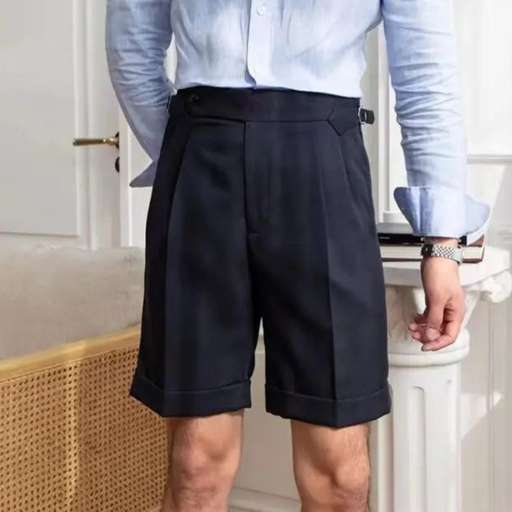 A person wearing a light blue button-up shirt tucked into high-waisted pleated shorts with a tailored fit, standing indoors near a light-colored cane chair, exudes an old money aesthetic.