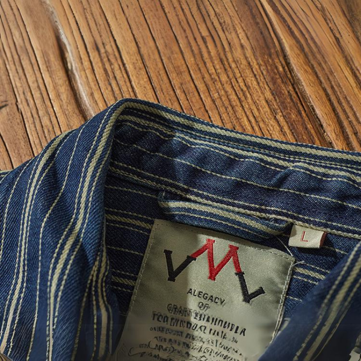 A close-up of a blue and white striped denim shirt collar with vertical stripes, featuring a fabric label inside marked with a red and black logo, the brand name ALEGACY, and a size L tag, laid on a wooden surface.