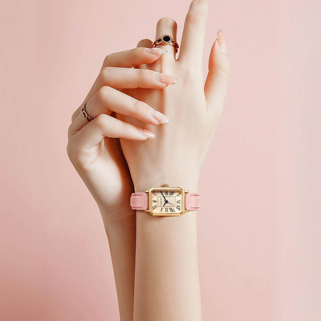 A hand with manicured nails wearing a gold rectangular face watch with a pink genuine leather strap is gently held by another hand, both set against a soft pink background.