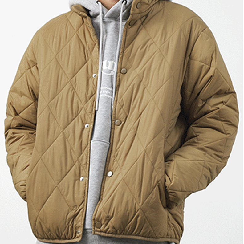 A person wearing a light brown, diamond-stitched pattern, quilted jacket over a grey hooded sweatshirt stands against a plain white background. Their hands are in the padded jacket pockets.