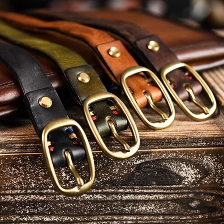 Four leather belts in black, green, brown, and dark brown with gold buckles are laid side by side on a wooden surface, showcasing country fashion style. The belts feature visible stitching near the buckles and metal rivets.