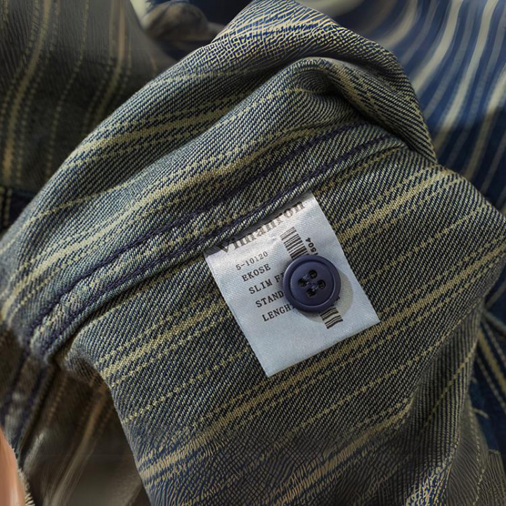 A close-up of a striped denim shirt with greenish and beige vertical stripes. A white label displays size and care instructions, and a dark blue button is sewn onto the relaxed-fit button-down fabric.