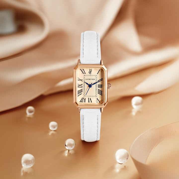 A rectangular face watch with a gold case, Roman numerals, and a genuine leather strap is displayed upright on a beige satin surface, surrounded by scattered pearls and soft, diffused lighting.