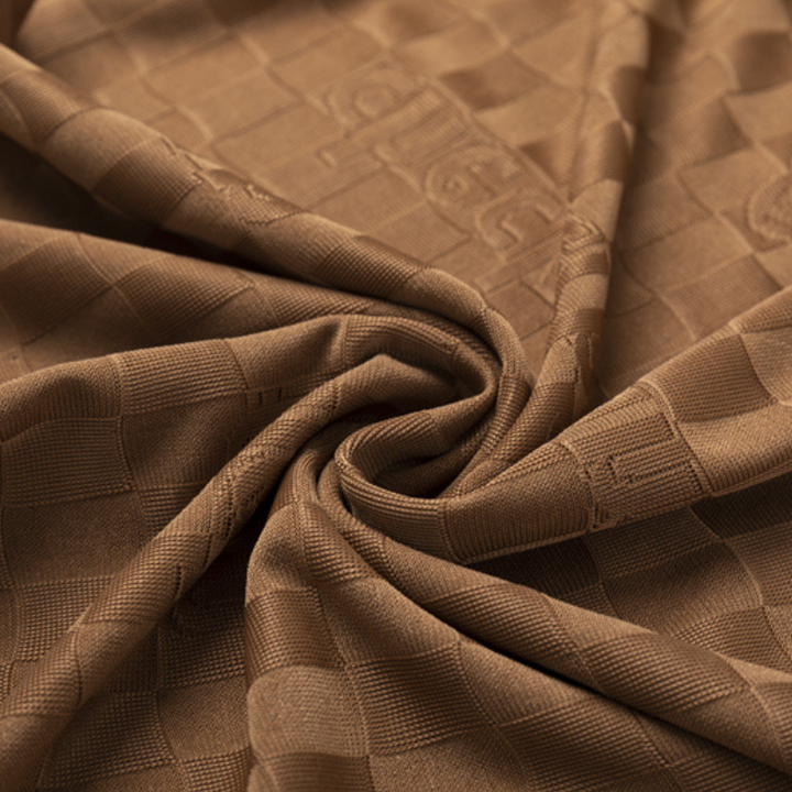 Close-up of brown Silk Cotton blend fabric with a subtle checkerboard pattern and embossed logo, gathered and twisted at the center to showcase its texture and weave details.