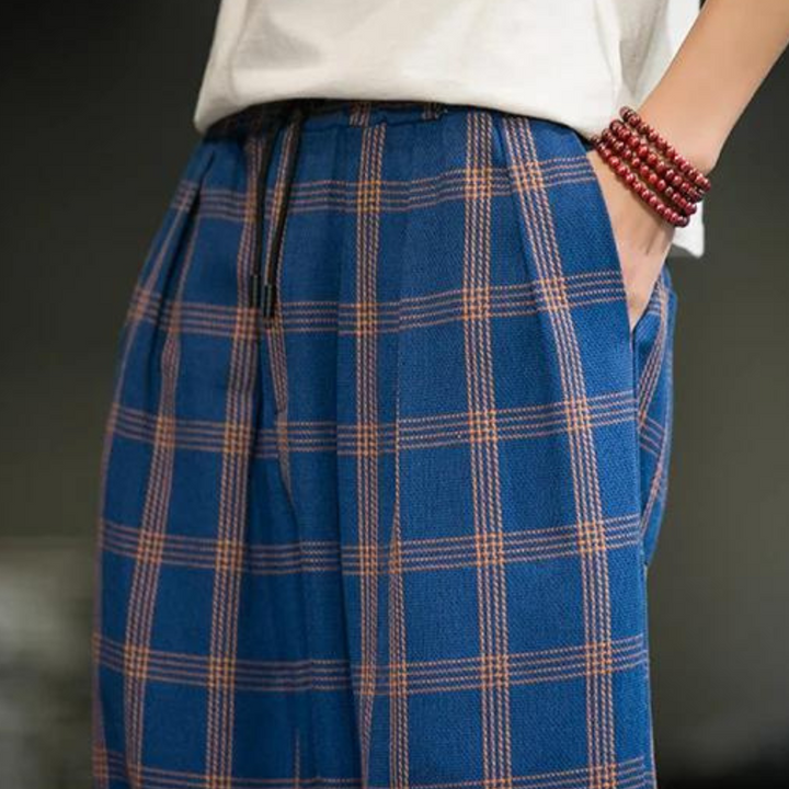 Person wearing blue plaid pants with orange lines—a bold fashion statement—paired with a white shirt and a red beaded bracelet, one hand in their pocket, shown from the waist down.