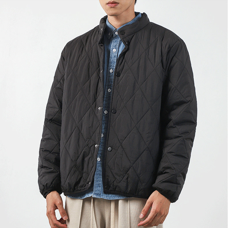 A person wearing a black, diamond-stitched padded jacket over a blue button-up shirt and beige pants stands against a plain white background. The jacket has a high collar and snap buttons. The persons face is partially out of frame.