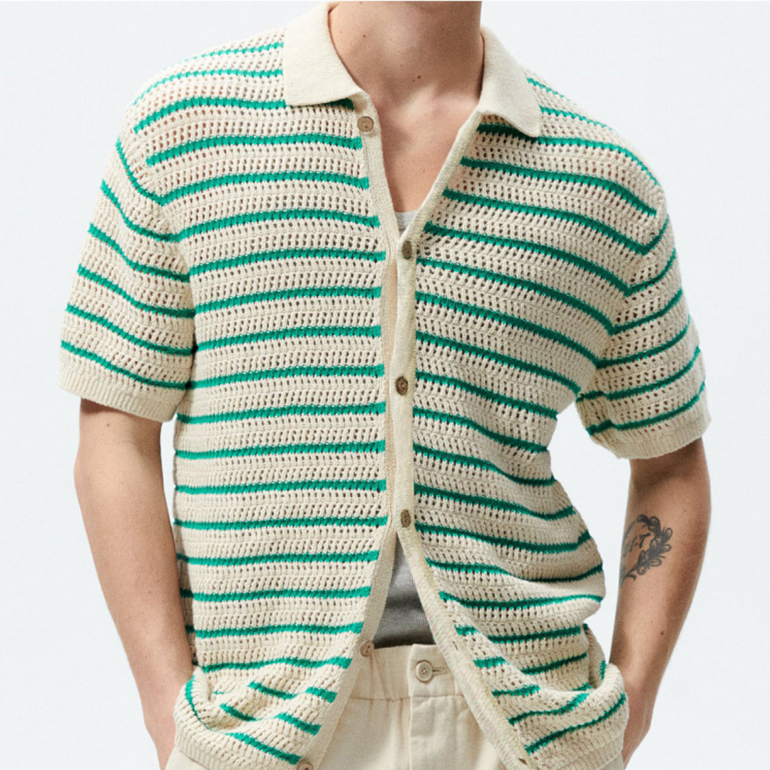 A person wears a cream and green striped open-knit design short-sleeve crochet shirt over a gray top, paired with cream pants. A visible arm tattoo adds to the streetwear vibe against a plain light background.