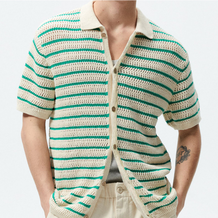 A person wears a cream and green striped open-knit design short-sleeve crochet shirt over a gray top, paired with cream pants. A visible arm tattoo adds to the streetwear vibe against a plain light background.