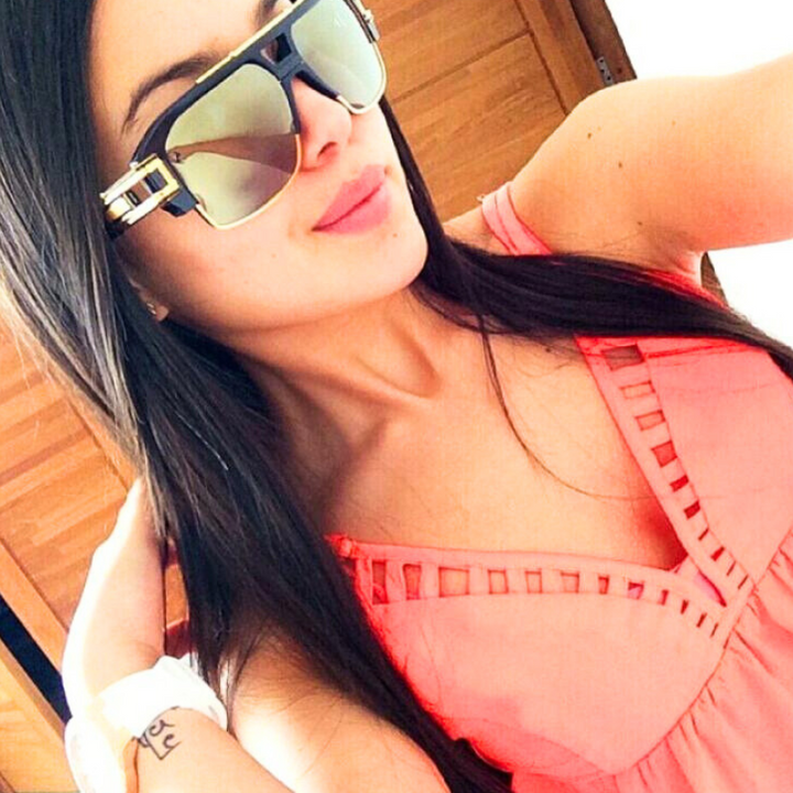 A woman with long dark hair takes a selfie in geometric sunglasses featuring UV400 protection. She wears a coral pink dress with cutout details and a white wristwatch, posing with one hand in her hair in front of wooden doors.
