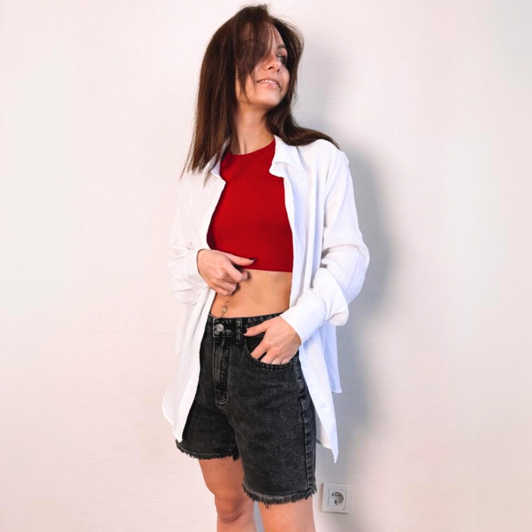 A woman stands against a plain light-colored wall, wearing a red crop top, a white unbuttoned shirt, and black wash denim shorts. She smiles and looks to the side, with one hand on her hip.