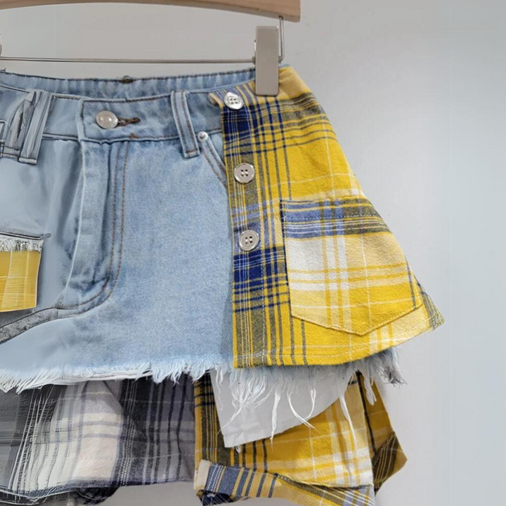 A close-up of an eclectic fashion patchwork skirt, combining blue denim and yellow plaid fabric with frayed edges, buttons, and visible stitching, hanging on a wooden hanger against a plain background.