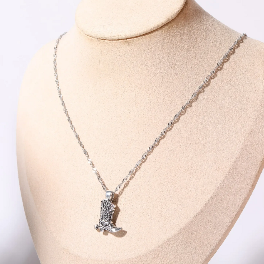 A silver cowgirl necklace featuring a decorative cowboy boot pendant is displayed on a beige jewelry bust, adding a touch of western jewelry charm.