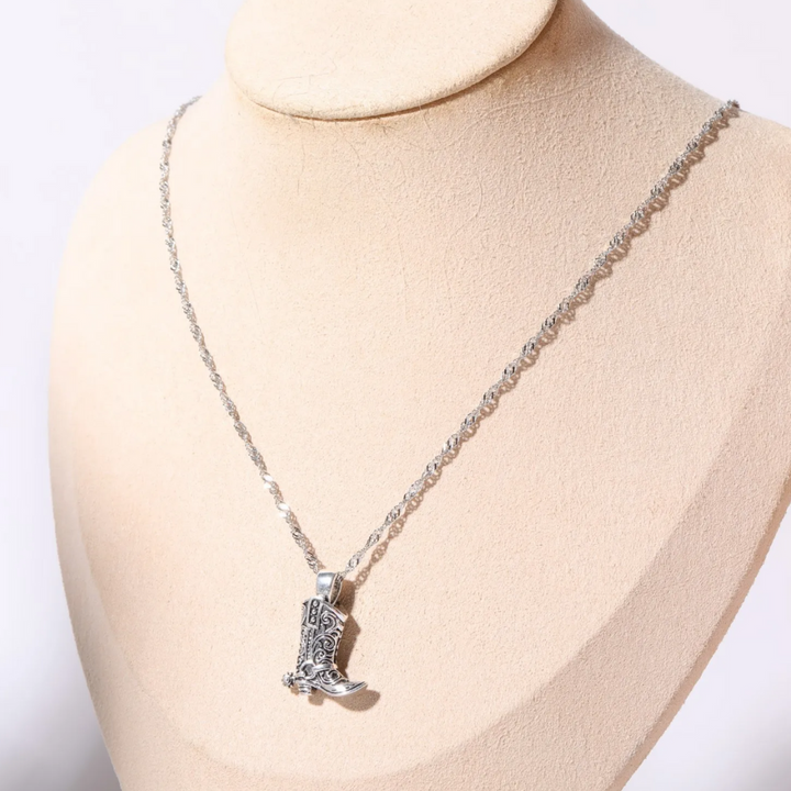 A silver cowgirl necklace featuring a decorative cowboy boot pendant is displayed on a beige jewelry bust, adding a touch of western jewelry charm.