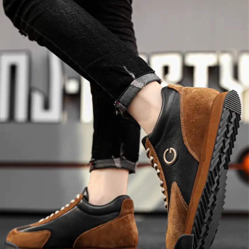 Person wearing luxe leather suede, brown and black sleek sneakers with a power button symbol on the side, standing on tiptoe against a blurred urban background. The person is wearing rolled-up, black jeans.