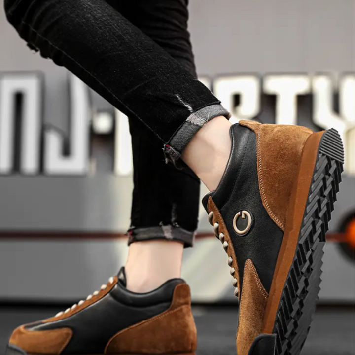 Person wearing luxe leather suede, brown and black sleek sneakers with a power button symbol on the side, standing on tiptoe against a blurred urban background. The person is wearing rolled-up, black jeans.