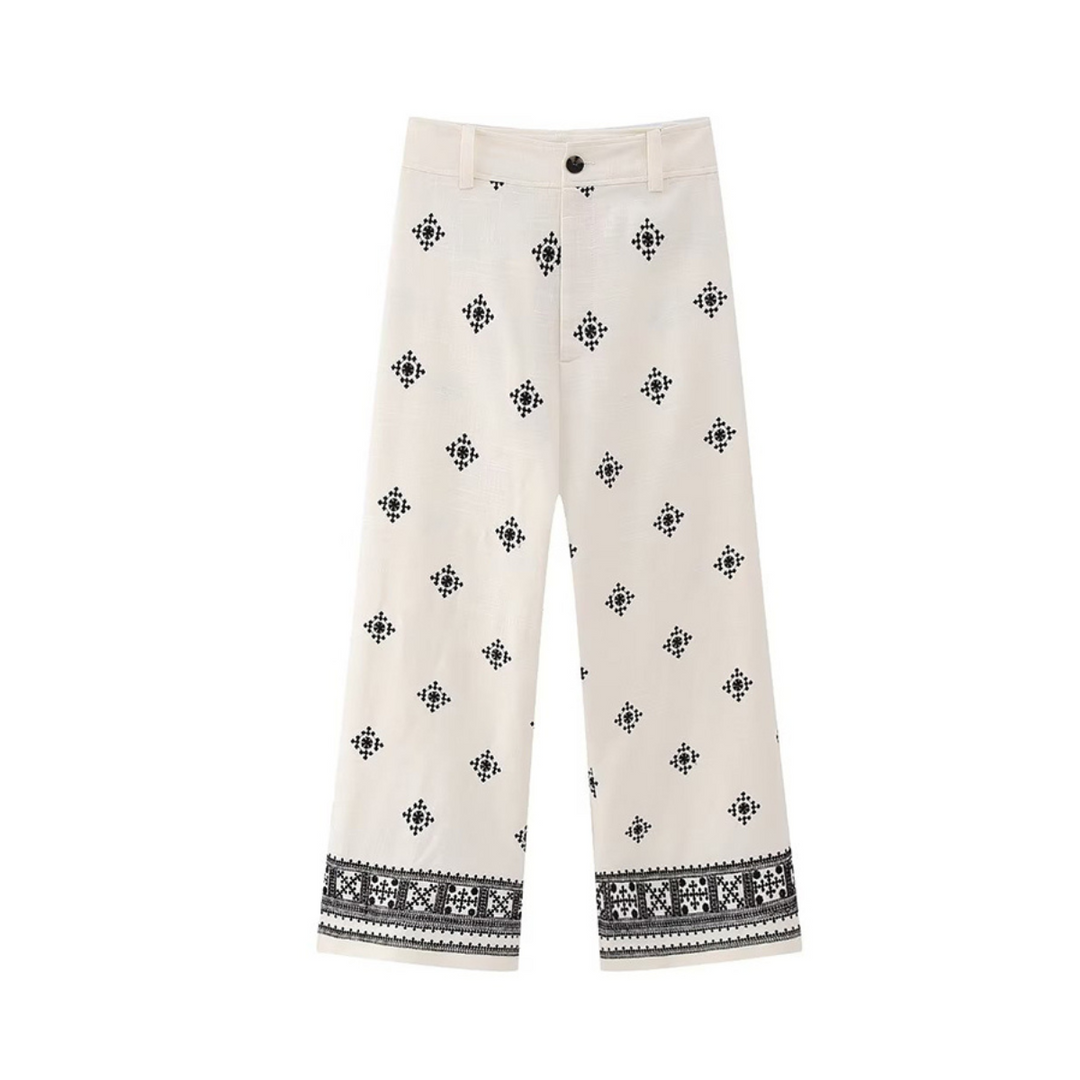 White wide-leg trousers with black geometric and floral embroidery scattered throughout and a decorative embroidered border near the hem. These boho chic pants feature a button and zip closure for easy wear.