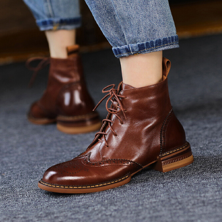 A person wearing rolled-up blue jeans and brown genuine leather lace-up ankle boots stands on a carpeted floor. The boots, with decorative stitching and a small heel, make a classic statement for the urban trendsetter.