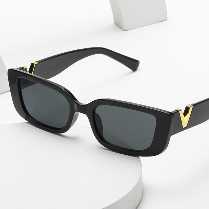 Black rectangular designer sunglasses with dark lenses and geometric frames, featuring gold V-shaped accents on the hinges. Displayed on a white surface with curved white props in the background. Offers UV400 protection.