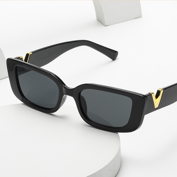 Black rectangular designer sunglasses with dark lenses and geometric frames, featuring gold V-shaped accents on the hinges. Displayed on a white surface with curved white props in the background. Offers UV400 protection.