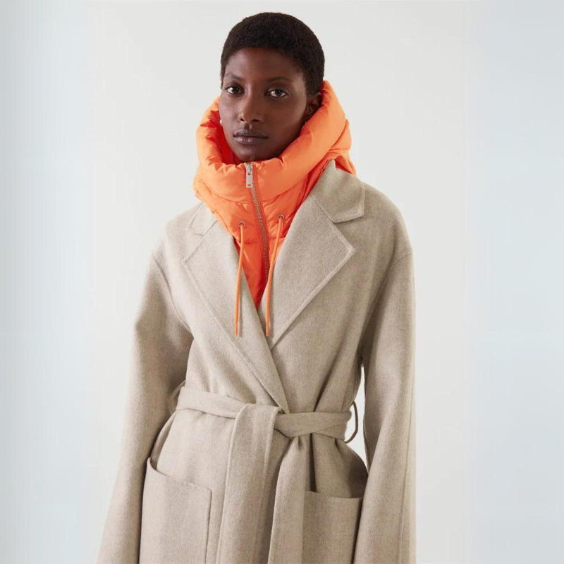 A person wearing a light beige belted coat over a lightweight orange zip-up puffer jacket with a high collar, standing against a plain light background.
