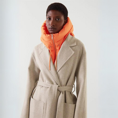 A person wearing a light beige belted coat over a lightweight orange zip-up puffer jacket with a high collar, standing against a plain light background.