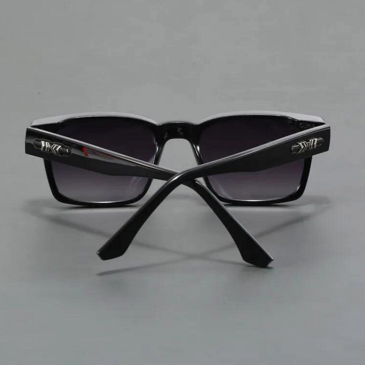 A pair of black sunglasses with dark lenses, shown from the back with the arms crossed. The durable frames feature metallic decorative details near the hinges. The background is a plain gray surface.