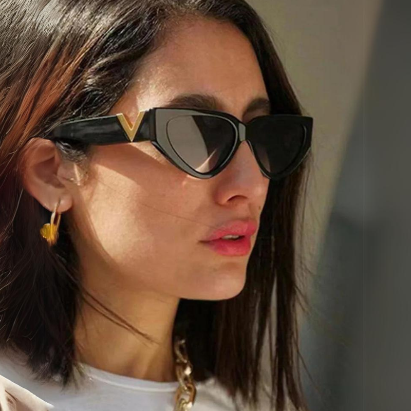 A woman with shoulder-length dark hair wears bold frame sunglasses with gold accents, gold hoop earrings, and a white top. She is looking slightly to the side. These vintage cat eye sunglasses add a chic touch to her look.