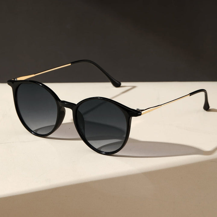 Round black sunglasses with dark lenses and thin gold metal arms, offering UV protection and anti-glare features, placed on a light surface with a dark background.