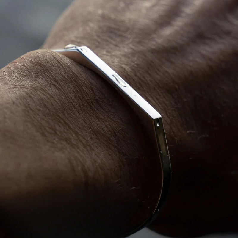 A close-up of a person’s wrist wearing a minimalist cuff in a sleek, modern design. The stainless steel bracelet showcases angular lines, while skin texture and light reflections on the metal are clearly visible.