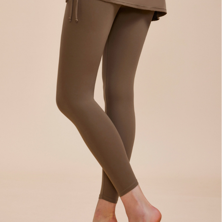 A person wearing taupe-colored high-waisted leggings and a matching skirt stands barefoot with one foot slightly raised against a beige background—an ideal look for an autumn wardrobe.