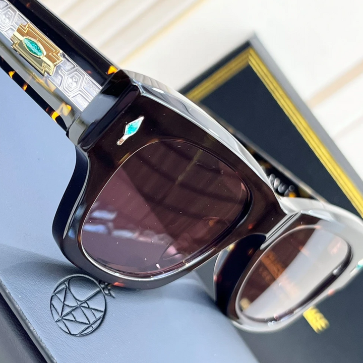 Close-up of stylish retro sunglasses with decorative gold and turquoise details on the arms, resting on a textured case with a geometric logo, and a black box with gold accents in the background. UV400 protection ensures both style and safety.