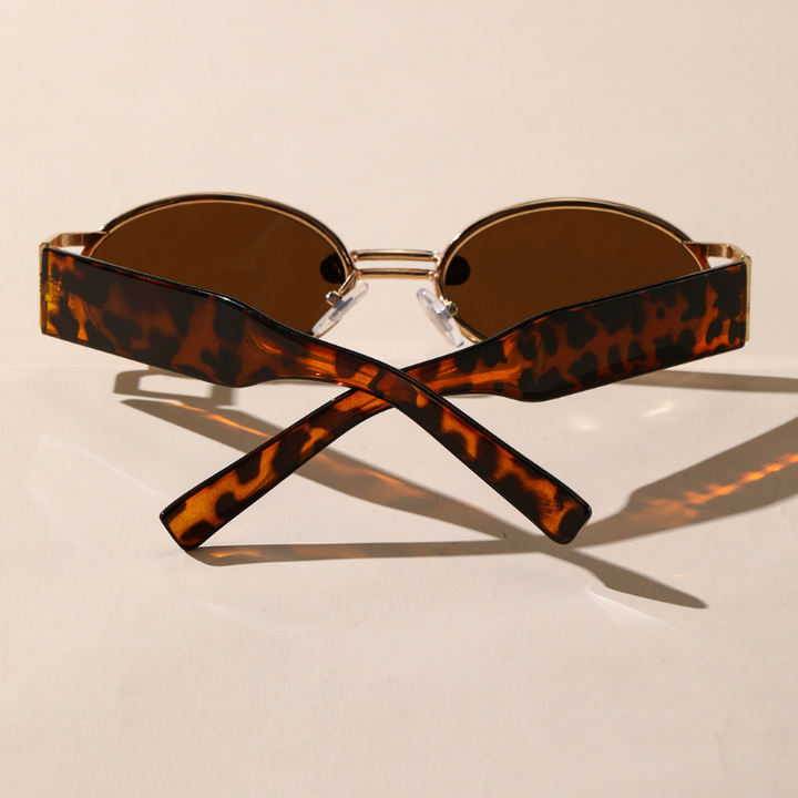 A pair of brown retro oval sunglasses with a durable metal frame and thick, tortoiseshell-patterned arms crossed at the ends, displayed on a light beige surface with soft shadows.
