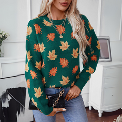 A woman with long blonde hair wears a comfortable green sweater with a maple leaf pattern in orange and yellow, light blue jeans, and holds a black purse with a gold chain. She stands indoors near white furniture and framed art.