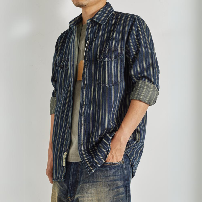 A man wears a relaxed-fit button-down with vertical stripes over a gray T-shirt and faded jeans, standing with one hand in his pocket. His face is partially out of the frame.