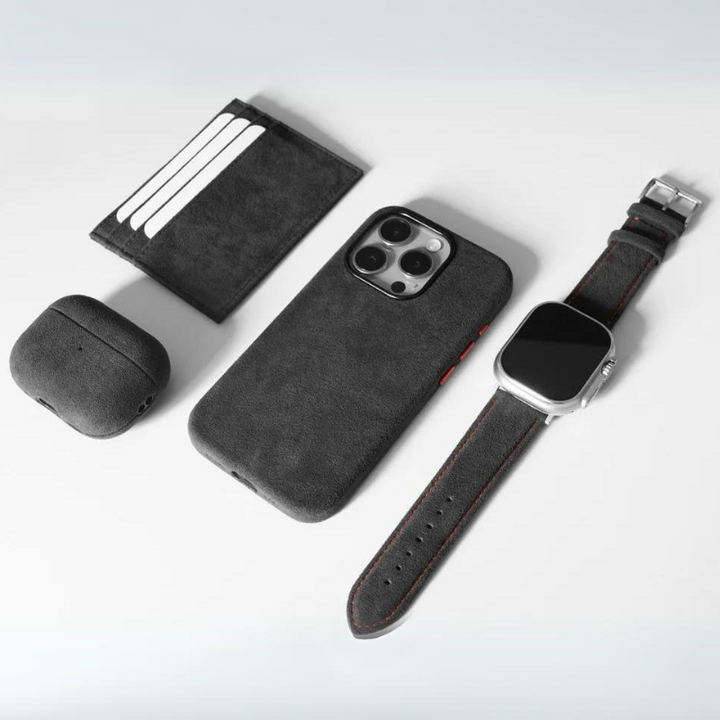 A flat lay of a black cardholder, smartphone case, wireless earbud case, and smartwatch with a black strap featuring a stainless steel buckle, all arranged on a light gray background.