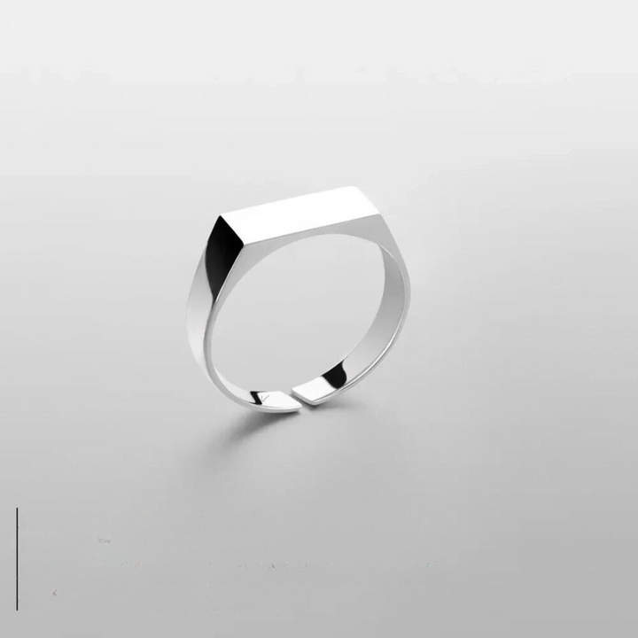 A minimalist stainless steel ring with a geometric, flat-topped design is displayed on a smooth, light gray surface. The ring casts a faint shadow, emphasizing its modern, scratch-resistant and sleek style.