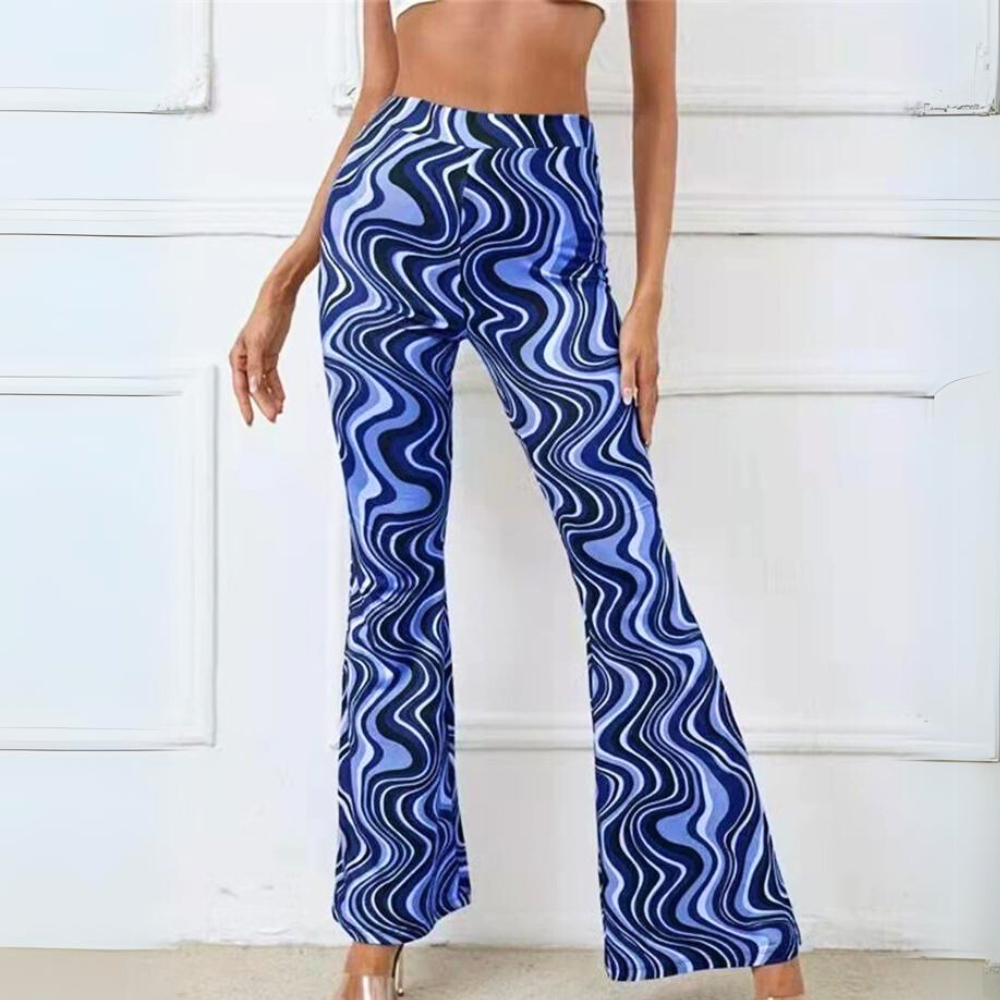 A woman wearing trendy statement pants—high-waisted, blue and white wavy-patterned psychedelic flare pants—stands in front of a white wall with decorative molding. Her face remains unseen in the image.