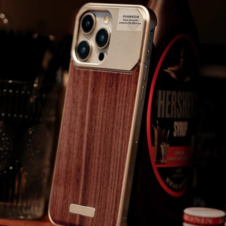 A smartphone with a stylish wood phone case stands upright on a surface. In the background, there is a bottle of Hershey’s chocolate syrup and a glass container with utensils.