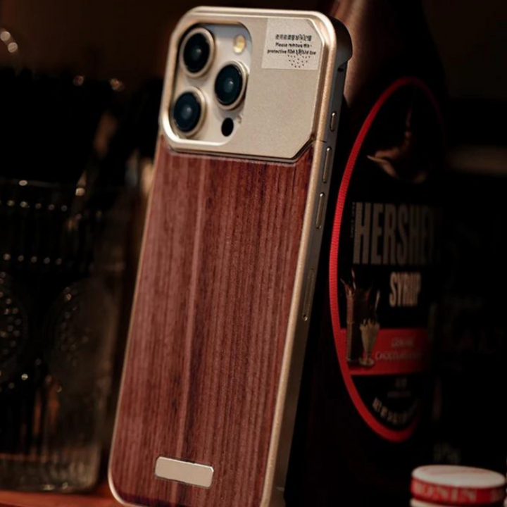 A smartphone with a stylish wood phone case stands upright on a surface. In the background, there is a bottle of Hershey’s chocolate syrup and a glass container with utensils.