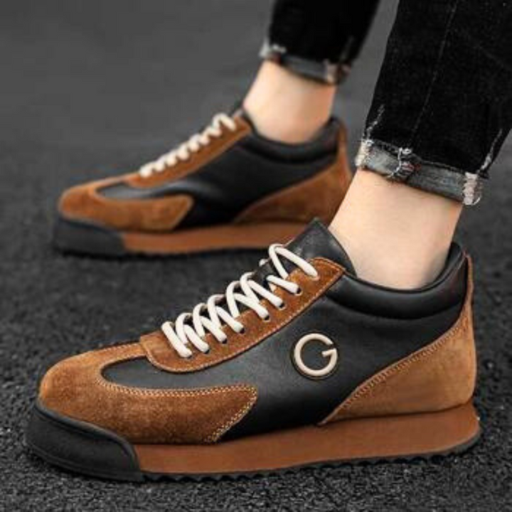 A person wearing luxe leather suede sneakers in black and brown with white laces and a G logo, standing on a dark textured surface. The sleek sneakers complement their black jeans with rolled cuffs, perfect for the urban professional.