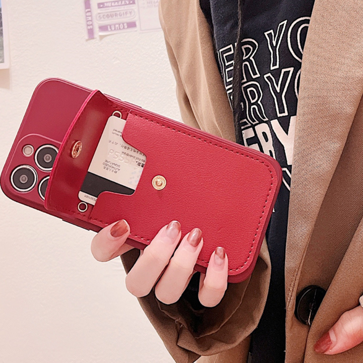 A person holds a phone with a red phone case wallet featuring a pocket as a secure cardholder. The person has painted nails and is wearing a brown coat and a black shirt with white text.