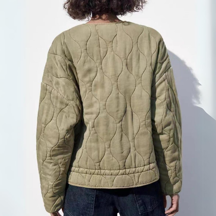 A person with curly hair, seen from behind, wears a light olive green quilted bomber with padded insulation and a wavy stitched pattern, paired with dark jeans, standing against a plain white background.