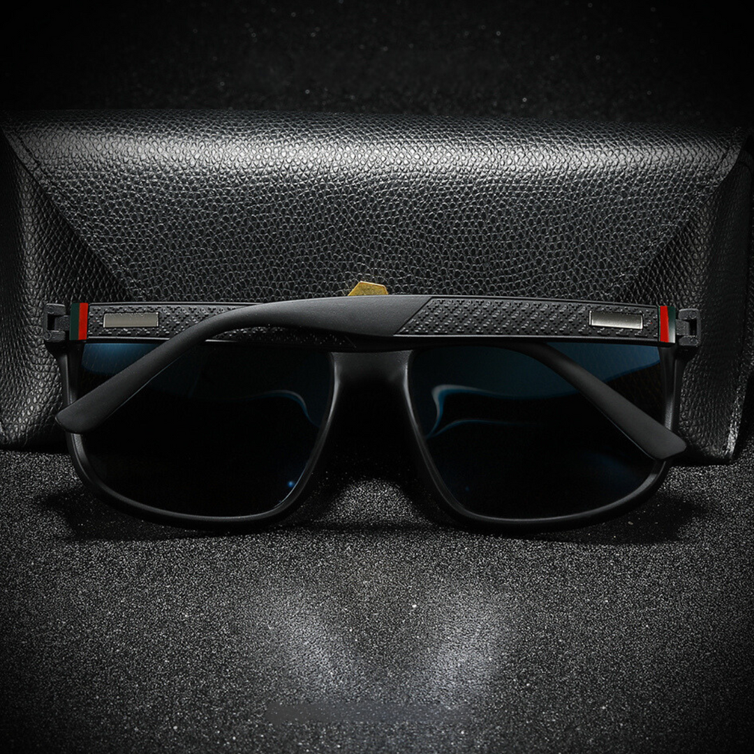 A pair of stylish black UV400 sunglasses with textured arms and a subtle red and green stripe detail rests in front of a closed black textured case on a dark, speckled surface.