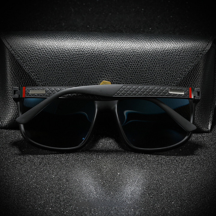A pair of stylish black UV400 sunglasses with textured arms and a subtle red and green stripe detail rests in front of a closed black textured case on a dark, speckled surface.