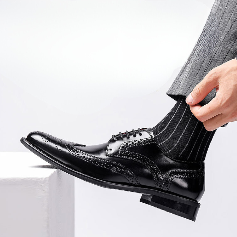 A person wearing a polished black premium leather brogue wingtip dress shoe with black pinstriped socks and gray suit pants rests their foot on a white surface.