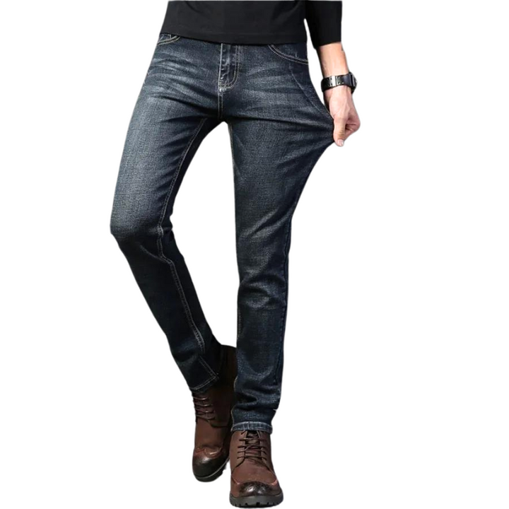 A person wearing dark blue stretch denim slim-fit jeans, a black long-sleeve shirt, brown lace-up boots, and a watch pulls on the side of the jeans to show their stretchiness. The background is white.