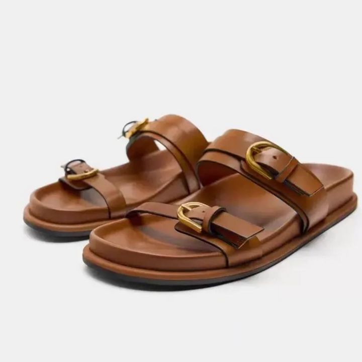 A pair of brown leather sandals with a leather upper and two straps, each featuring a large gold buckle, displayed on a white background.