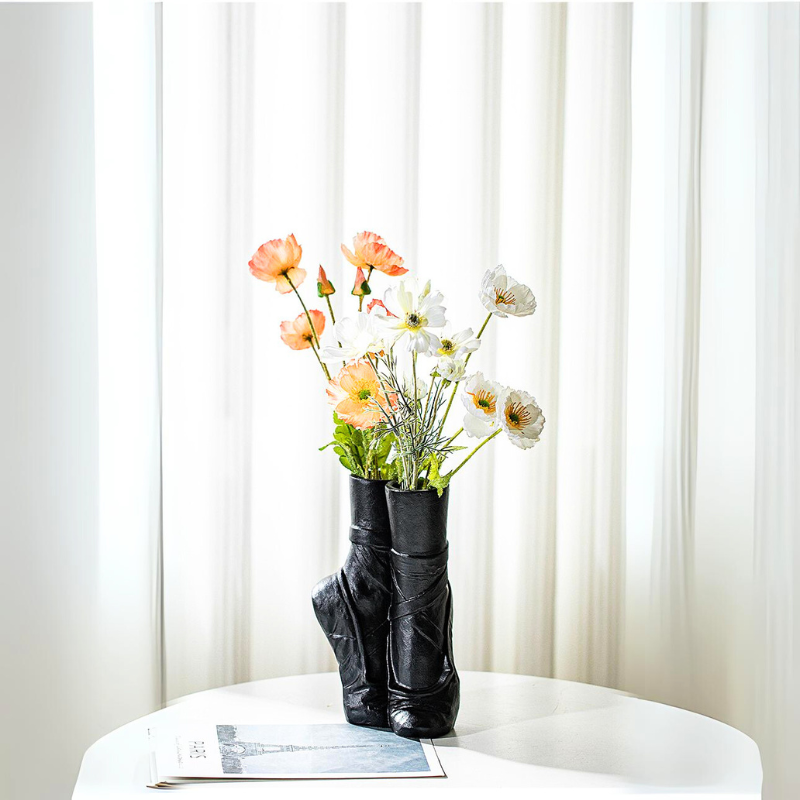 A black ballet-inspired ceramic vase shaped like pointe shoes holds orange and white flowers on a round white table, with a folded newspaper nearby and sheer white curtains in the background—perfect for contemporary chic décor.