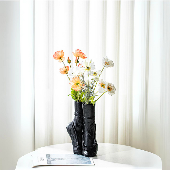 A black ballet-inspired ceramic vase shaped like pointe shoes holds orange and white flowers on a round white table, with a folded newspaper nearby and sheer white curtains in the background—perfect for contemporary chic décor.